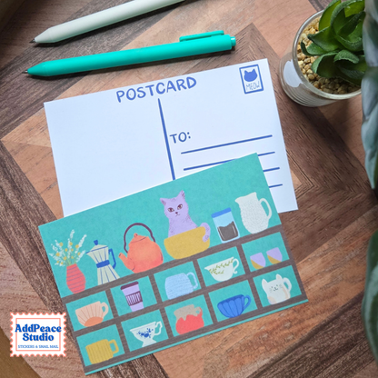 A postcard set displayed on a wooden shelf, featuring a white postcard with blue lines and a colorful illustrated postcard with tea-themed illustrations including a purple cat, teapot, cups, and mugs. A green pen, a white stylus, and a small potted plant are arranged around it.