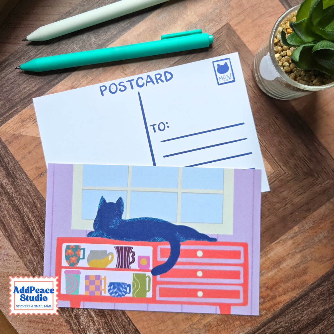 A postcard set displayed on a wooden tabletop, featuring a white postcard with blue printed lines and a colorful illustrated postcard showing a black cat lying on a red dresser with coffee mugs.