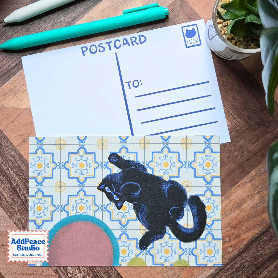 A postcard set displayed on a wooden tabletop, featuring a white postcard with blue writing lines and a patterned postcard with a black cat illustration and blue geometric tile design. A teal pen, succulent plant, and AddPeace Studio logo are also visible.