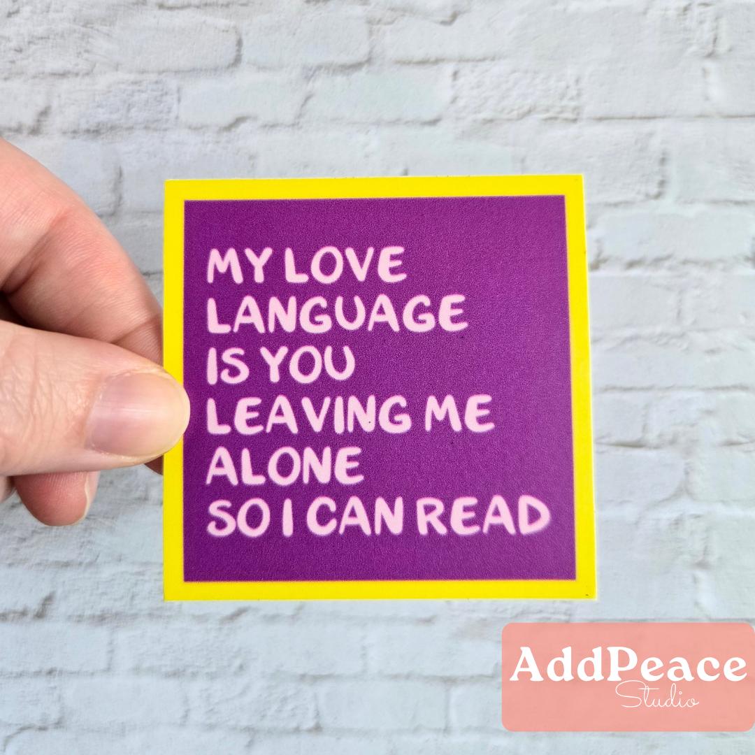 Hand holding a purple sticker with humorous text My love language is you leaving me alone so I can read against a light gray brick wall.