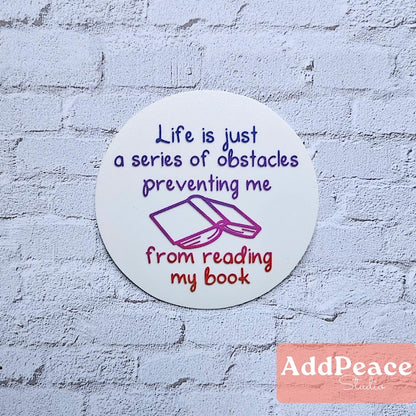 Circular sticker with Life is a series of obstacles preventing me from reading my book on a textured wall, featuring the brand 'AddPeace'