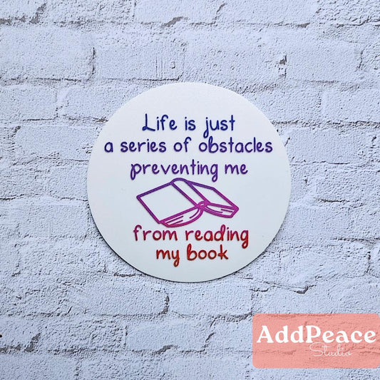 Circular sticker with Life is a series of obstacles preventing me from reading my book on a textured wall, featuring the brand 'AddPeace'