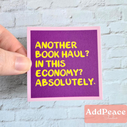 Hand holding a purple sticker with yellow text on a light gray brick wall background, featuring the brand 'AddPeace Studios'.
