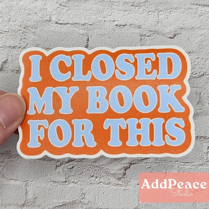 Sticker with 'I Closed My Book For This' text held by a hand on a gray textured background, with 'AddPeace' branding.