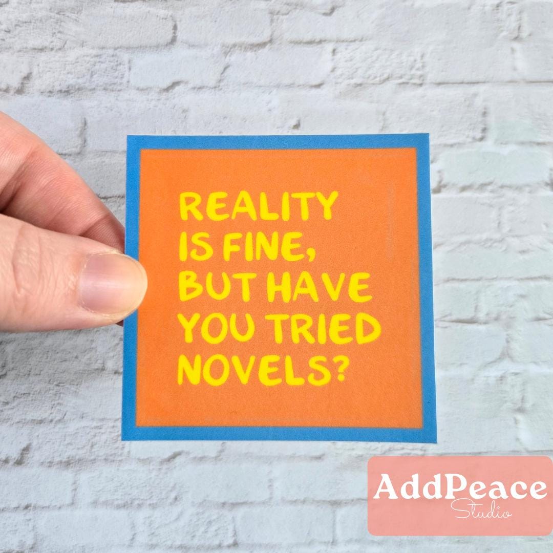 Hand holding a square sticker with text 'Reality is fine, but have you tried novels?' against a light gray brick wall.