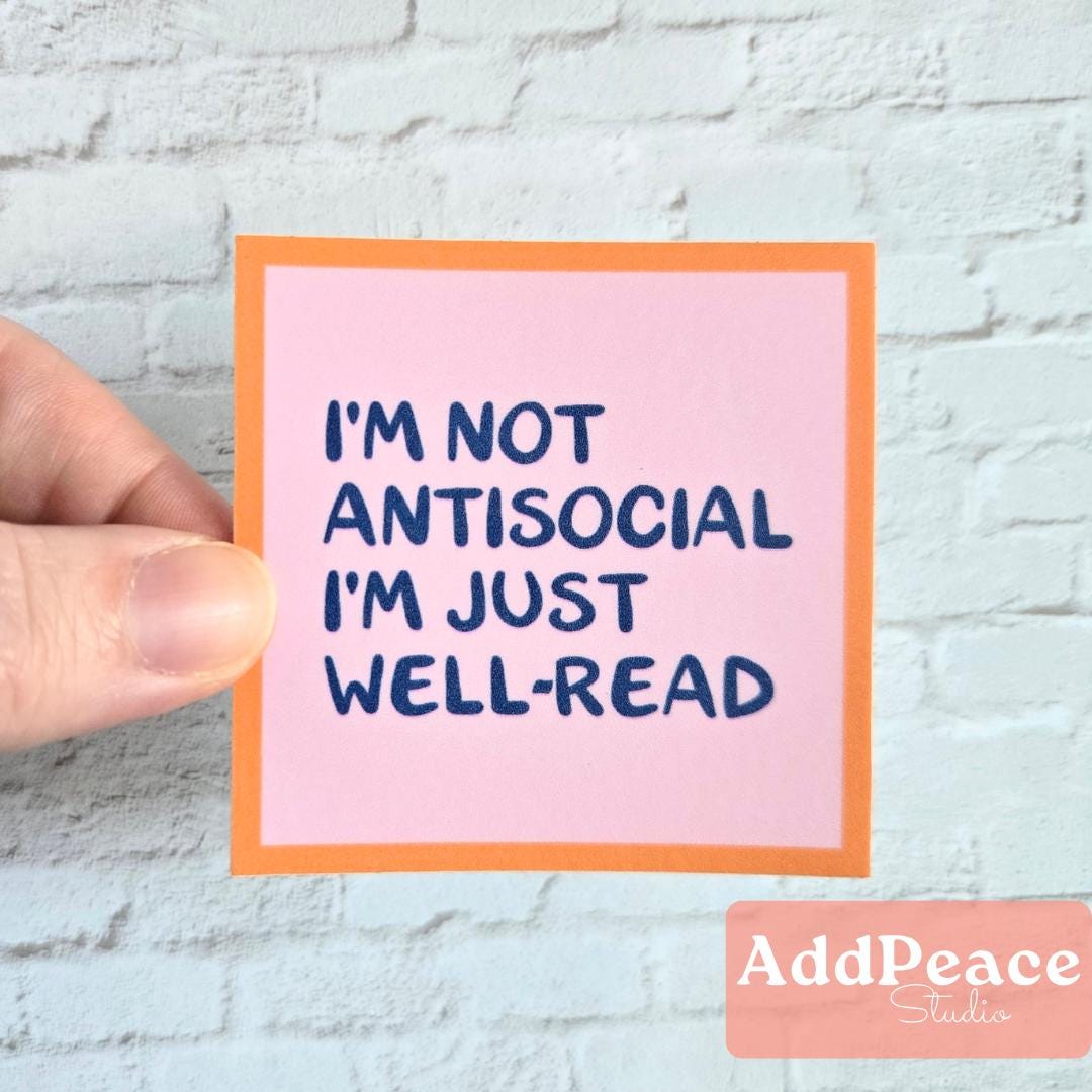 Hand holding a pink sticker with text 'I'm not antisocial, I'm just well-read' against a light gray brick wall.