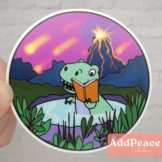 Sticker with a cartoon dinosaur reading a book in front of a volcano, held by a hand.