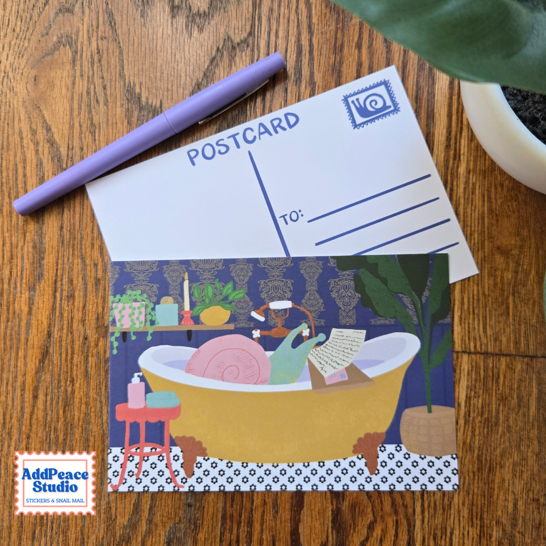 A postcard with a whimsical illustration of a snail relaxing in a yellow bathtub reading mail, styled on a wooden tabletop next to a purple pen and a plant.