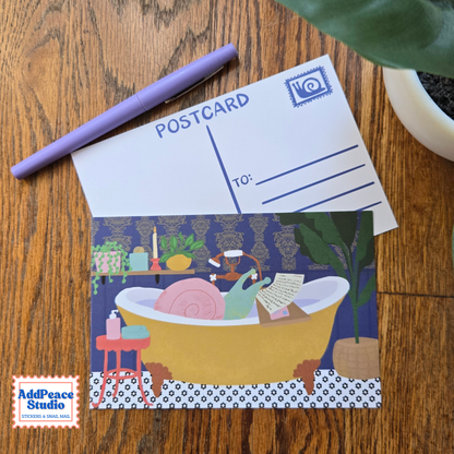 A postcard with a whimsical illustration of a snail relaxing in a yellow bathtub reading mail, styled on a wooden tabletop next to a purple pen and a plant.