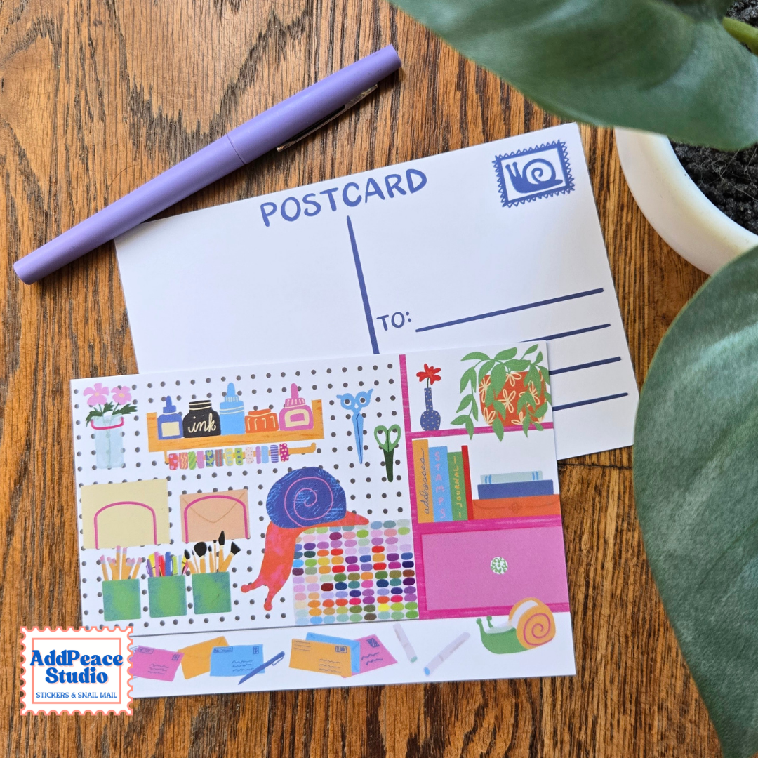 A postcard stationery set displayed on a wooden desk, featuring a white postcard with blue writing lines and a colorful illustrated desk-themed postcard featuring a curious snail. A purple pen and green plant leaves are styled around it, with an 'AddPeace Studio' brand logo in the bottom-left corner.