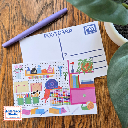 A postcard stationery set displayed on a wooden desk, featuring a white postcard with blue writing lines and a colorful illustrated desk-themed postcard featuring a curious snail. A purple pen and green plant leaves are styled around it, with an 'AddPeace Studio' brand logo in the bottom-left corner.