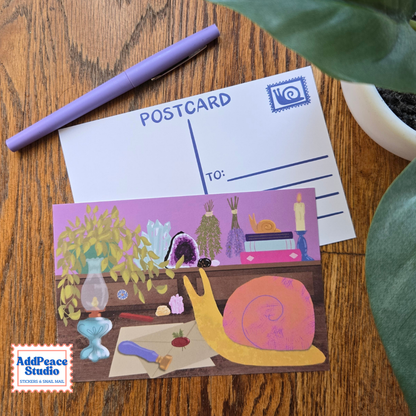 A postcard featuring whimsical illustrated artwork of a yellow snail with an orange shell, sealing a letter with wax, surrounded by plants, an oil lamp, and small objects on a wooden tabletop. A plain postcard back with writing lines and a postage stamp icon is placed behind it, along with a purple pen and green plant leaves.