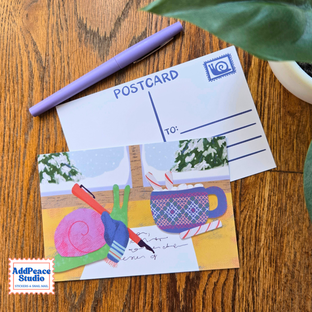 A handmade postcard featuring a colorful winter illustration of a green snail writing a letter beside a blue mug, displayed on a wooden table next to a purple pen and a blank postcard template. The 'AddPeace Studio' logo is visible in the bottom-left corner.