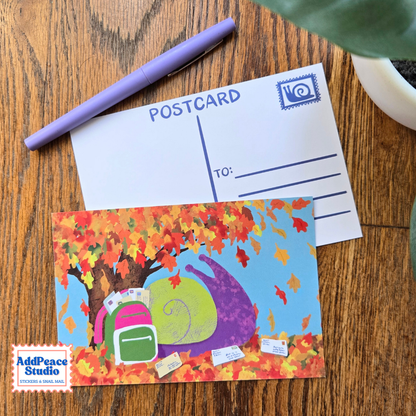 A colorful autumn-themed postcard featuring a purple snail under orange and red fall leaves, displayed on a wooden tabletop next to a purple pen and a white postcard.