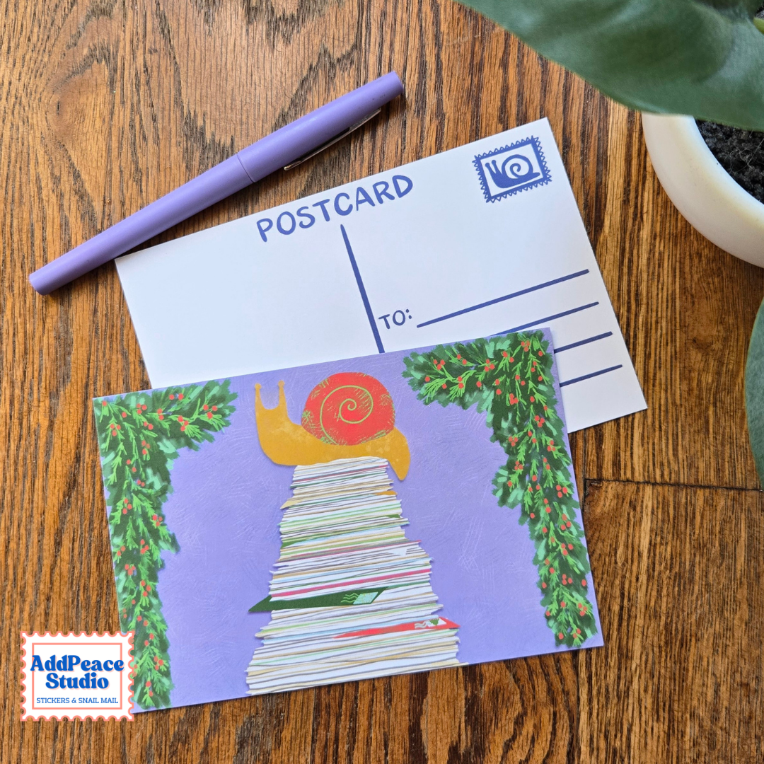 A postcard featuring an illustration of a yellow snail with a red shell climbing a stack of letters framed by green ivy, displayed on a wooden table next to a purple pen and a blank postcard. The AddPeace Studio logo is visible in the bottom-left corner.