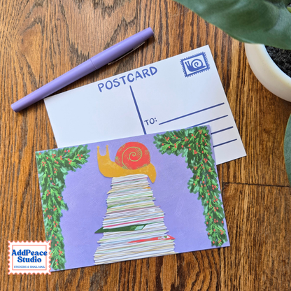 A postcard featuring an illustration of a yellow snail with a red shell climbing a stack of letters framed by green ivy, displayed on a wooden table next to a purple pen and a blank postcard. The AddPeace Studio logo is visible in the bottom-left corner.