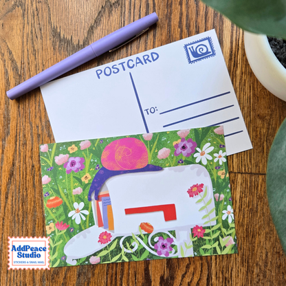 A postcard displayed on a wooden tabletop, featuring a white postcard back with blue printed lines and a stamp icon, layered over a colorful illustrated postcard with a mailbox, flowers, and a snail on top. A lavender pen rests above the cards, with a plant leaf partially visible at the right edge.