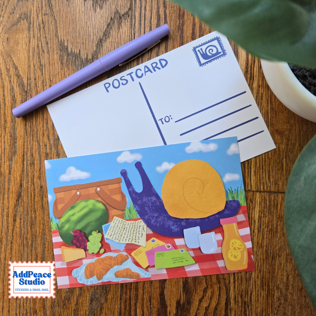 A colorful illustrated postcard featuring a snail picnic scene is placed on a wooden surface, with a white postcard back and a purple pen nearby. A plant leaf and a small pot are partially visible around the edges.
