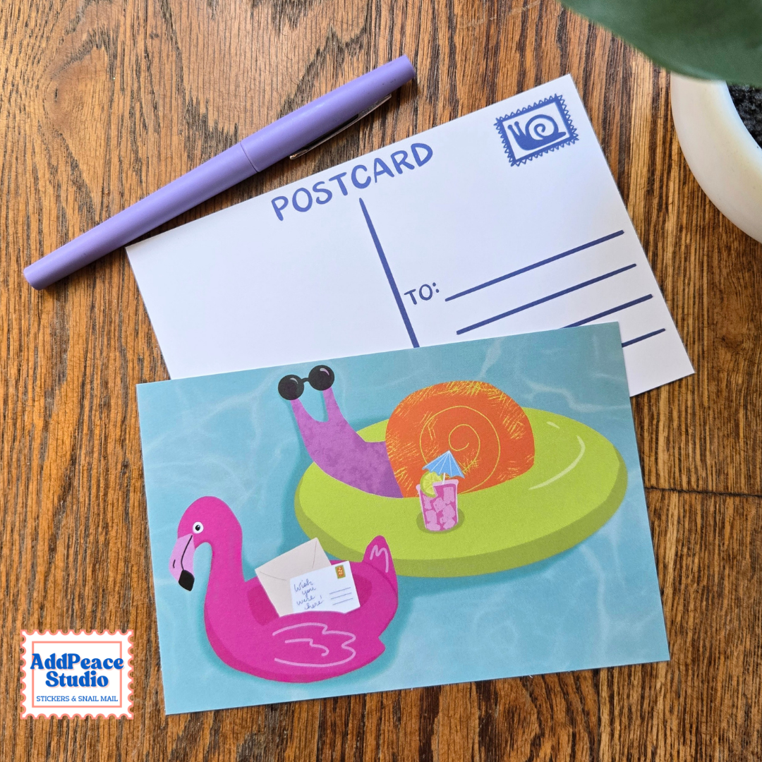 A postcard featuring a colorful illustration of a snail relaxing on a green float in a pool beside a pink flamingo float, displayed on a wooden table next to a purple pen and another postcard back.
