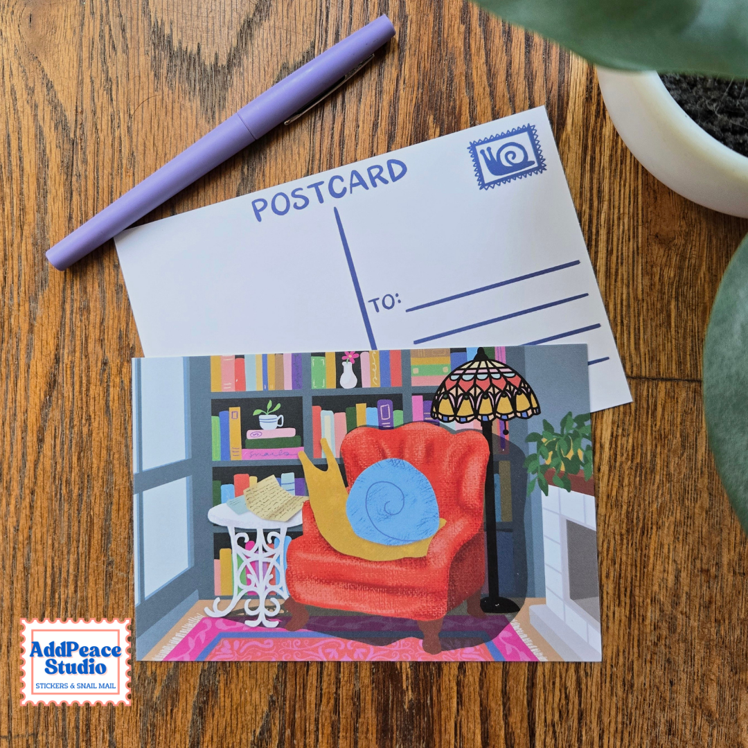 A postcard featuring a colorful illustration of a snail in a cozy living room with a red armchair, bookshelves, and a lamp, displayed on a wooden table next to a purple pen and a plant. The back of the postcard is also visible.