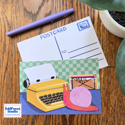 A postcard featuring a colorful illustration of a yellow typewriter and a pink snail on a green checkered background, displayed on a wooden desk with a purple pen and a plant.